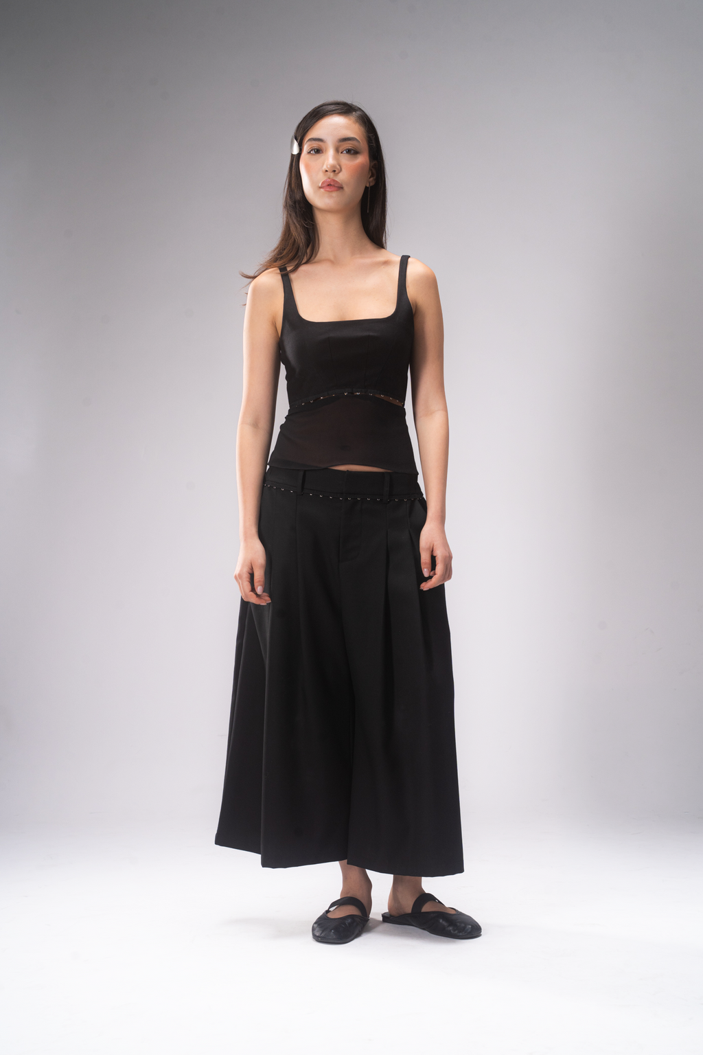 A woman stands against a light gray studio background wearing a black sleeveless top with a sheer mesh panel and a long black skirt with a studded waistband, paired with black flats.