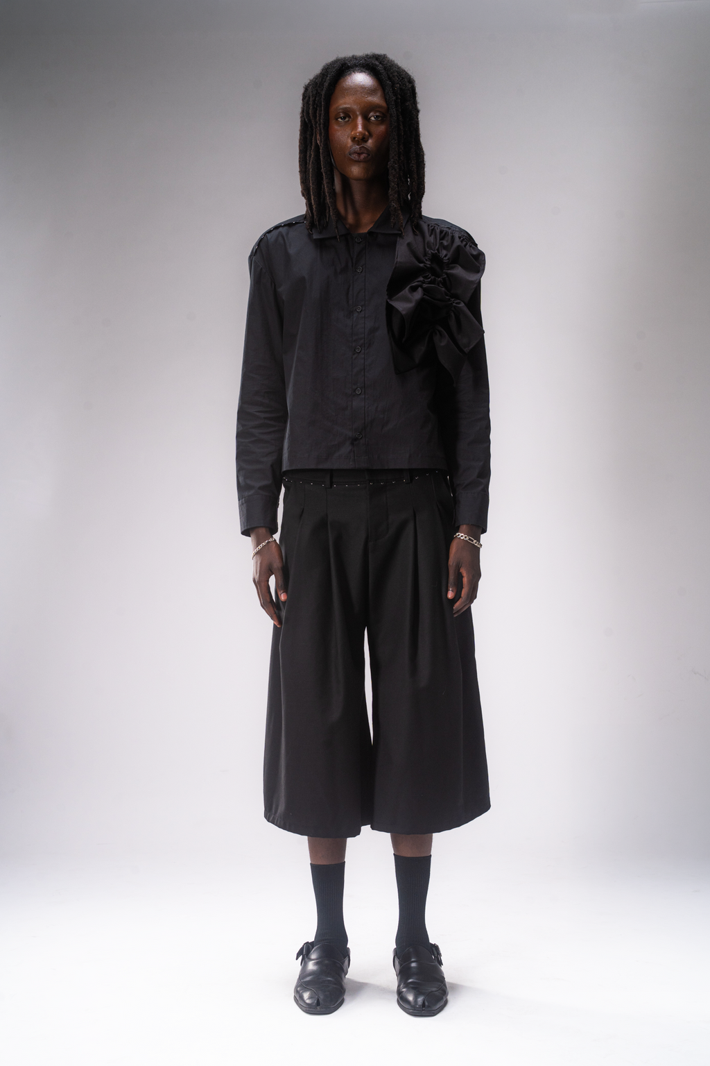 A model stands against a light gray studio background wearing a black long-sleeve button-up shirt with a large ruffled shoulder detail, black pleated cropped wide-leg pants, black socks, and black shoes.