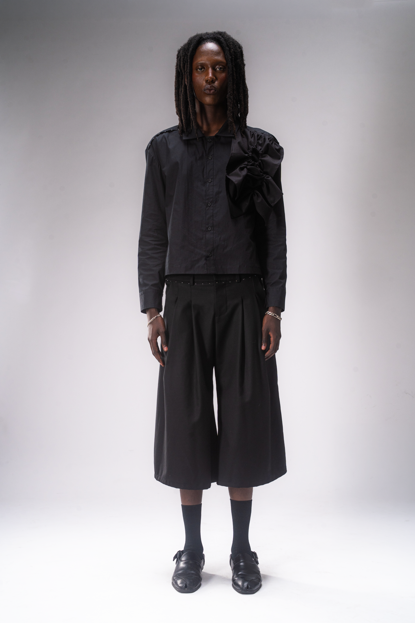 A model stands against a light gray studio background wearing a black long-sleeve button-up shirt with a large ruffled shoulder detail, black pleated cropped wide-leg pants, black socks, and black shoes.