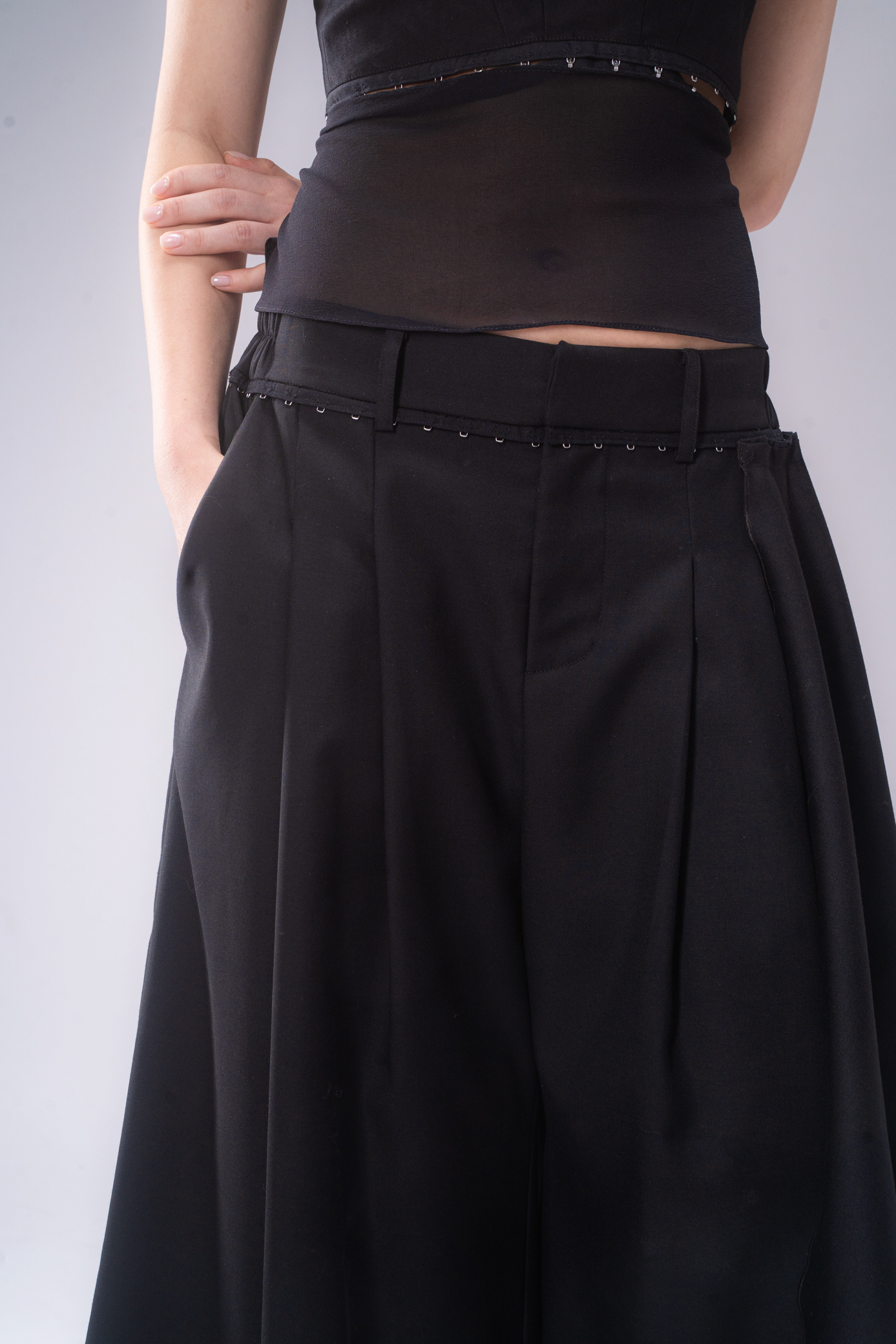 A close-up of a model wearing black wide-leg pleated trousers set against a plain light gray background.