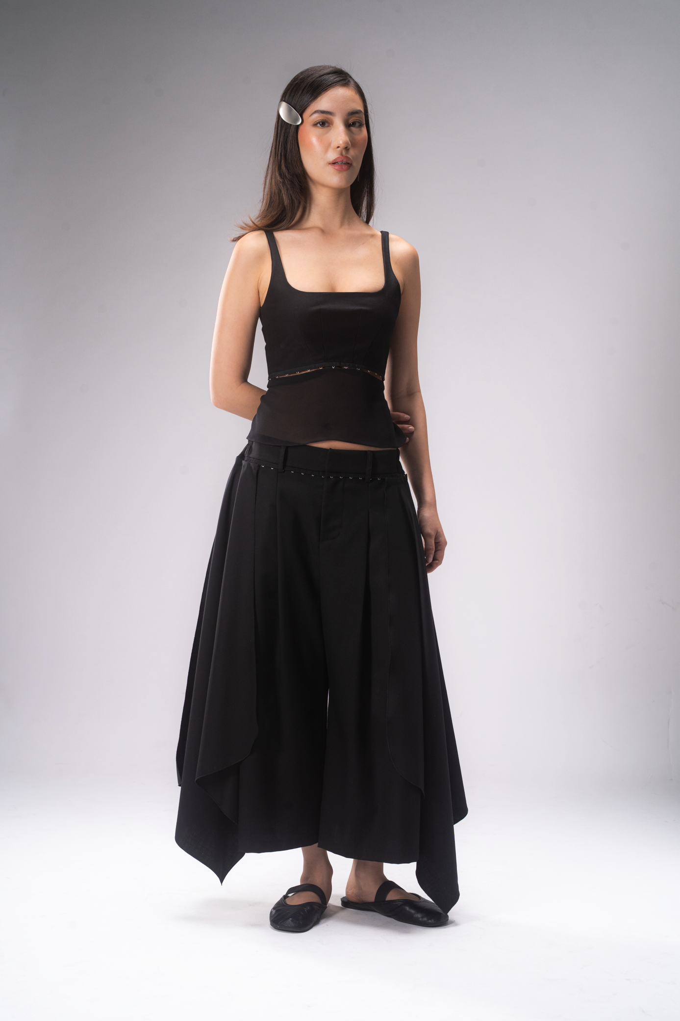 A female model stands against a light grey studio background wearing a black tank top and pleated cropped pants with removable wing panels