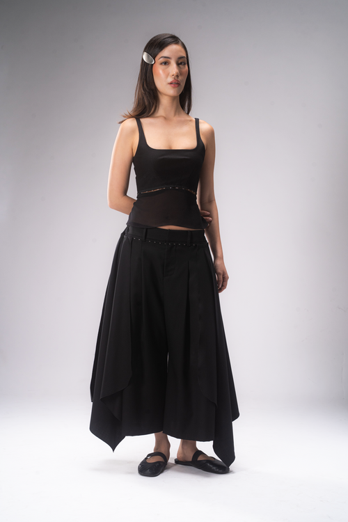 A female model stands against a light grey studio background wearing a black tank top and pleated cropped pants with removable wing panels