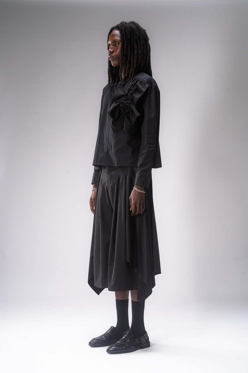 A male model stands against a light grey studio background wearing a black shirt and pleated cropped pants with removable wing panels